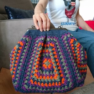 K&ouml;nnte beinhalten: Handgefertigte H&auml;keltasche im Granny-Square-Muster. Die Tasche hat eine dunkelblaue Grundfarbe mit leuchtenden Quadraten in Orange, Lila und Gr&uuml;n. Sie hat einen Metallverschluss und eine strukturierte Form, ideal f&uuml;r den Transport von Essentials.