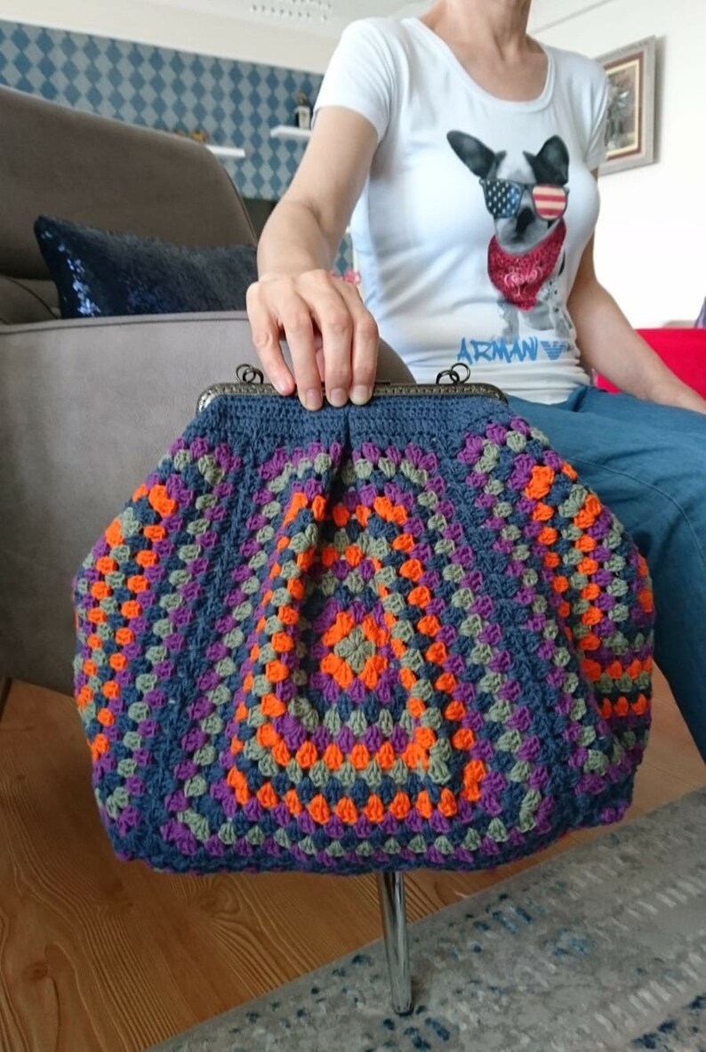 K&ouml;nnte beinhalten: Handgefertigte H&auml;keltasche im Granny-Square-Muster. Die Tasche hat eine dunkelblaue Grundfarbe mit leuchtenden Quadraten in Orange, Lila und Gr&uuml;n. Sie hat einen Metallverschluss und eine strukturierte Form, ideal f&uuml;r den Transport von Essentials.