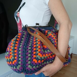 K&ouml;nnte beinhalten: Handgefertigte H&auml;keltasche mit farbenfrohem Granny-Square-Muster in Blau-, Orange-, Lila- und Gr&uuml;nt&ouml;nen. Die Tasche hat einen braunen Lederriemen und einen Metallverschluss. Ein stilvolles und einzigartiges Accessoire.