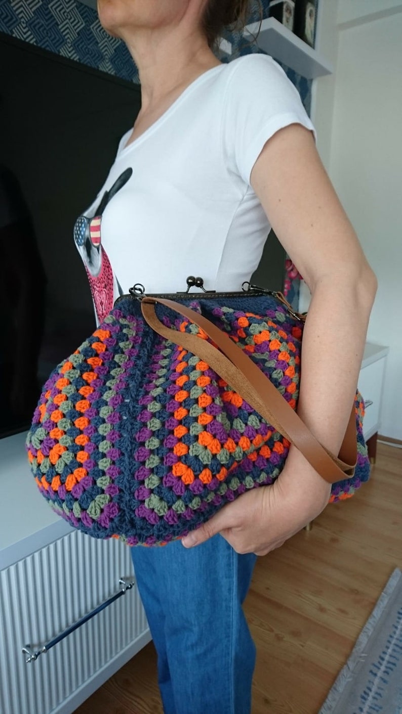 K&ouml;nnte beinhalten: Handgefertigte H&auml;keltasche mit farbenfrohem Granny-Square-Muster in Blau-, Orange-, Lila- und Gr&uuml;nt&ouml;nen. Die Tasche hat einen braunen Lederriemen und einen Metallverschluss. Ein stilvolles und einzigartiges Accessoire.