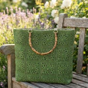 May include: A sage green crochet tote bag constructed from a grid of square floral motifs. The bag features two rounded bamboo handles attached with silver-toned metal rings, providing a structured opening for the rectangular textile body.