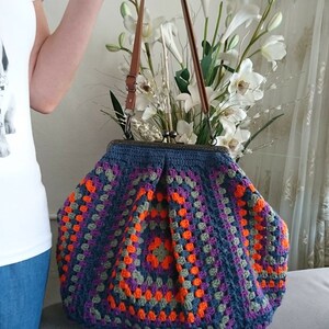 K&ouml;nnte beinhalten: Handgefertigte H&auml;keltasche mit farbenfrohem Granny-Square-Design. Die Tasche hat eine dunkelblaue Basis mit orangefarbenen, lila und grauen Quadraten. Sie hat braune Ledergriffe und einen Metallverschluss, ideal f&uuml;r den Alltag.