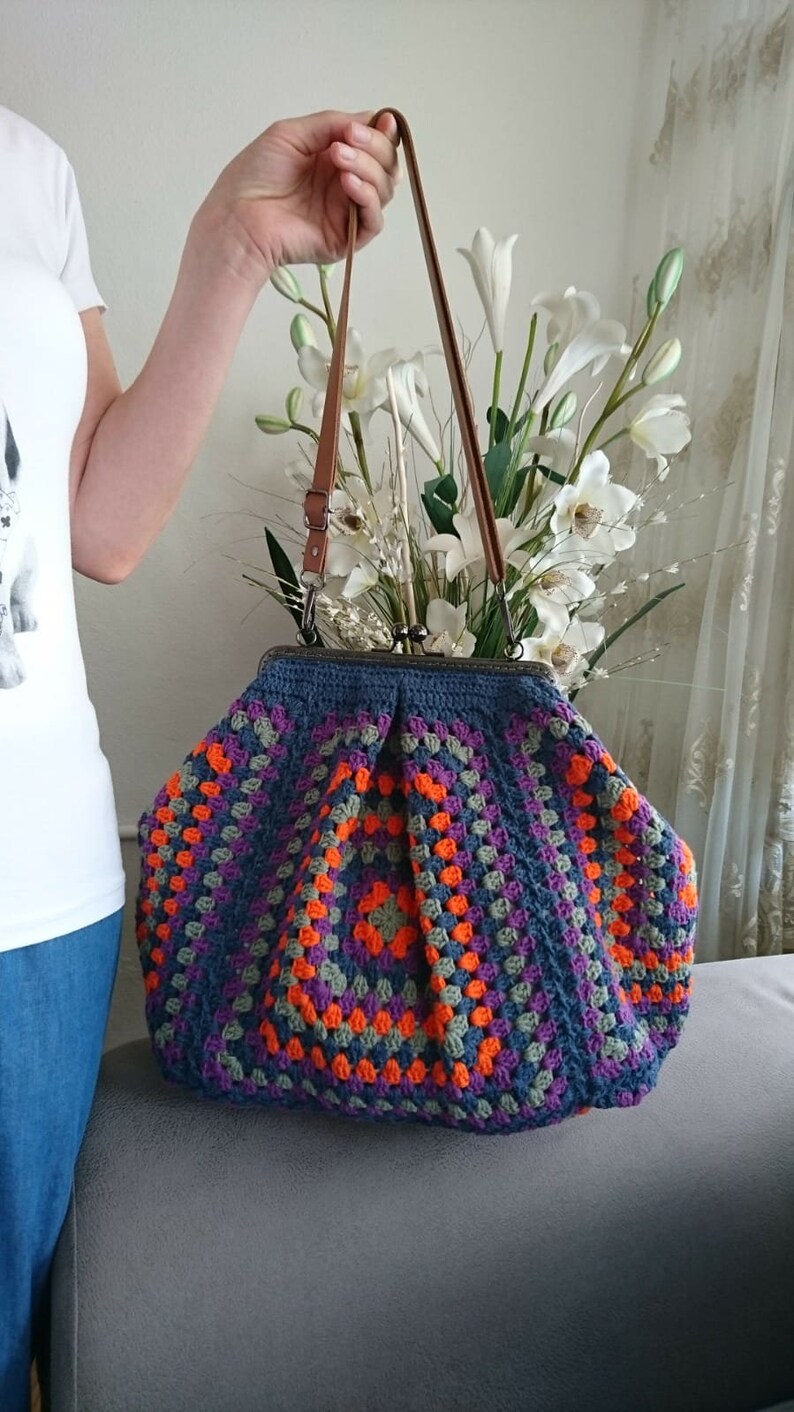K&ouml;nnte beinhalten: Handgefertigte H&auml;keltasche mit farbenfrohem Granny-Square-Design. Die Tasche hat eine dunkelblaue Basis mit orangefarbenen, lila und grauen Quadraten. Sie hat braune Ledergriffe und einen Metallverschluss, ideal f&uuml;r den Alltag.