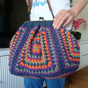 K&ouml;nnte beinhalten: Handgefertigte H&auml;keltasche im Granny-Square-Muster. Die Tasche hat eine dunkelblaue Basis mit orangefarbenen, lila und gr&uuml;nen Quadraten. Sie hat einen Metallverschluss und einen braunen Leder-Schulterriemen.