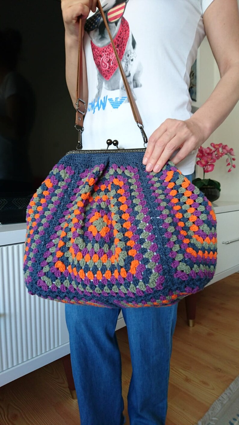K&ouml;nnte beinhalten: Handgefertigte H&auml;keltasche im Granny-Square-Muster. Die Tasche hat eine dunkelblaue Basis mit orangefarbenen, lila und gr&uuml;nen Quadraten. Sie hat einen Metallverschluss und einen braunen Leder-Schulterriemen.