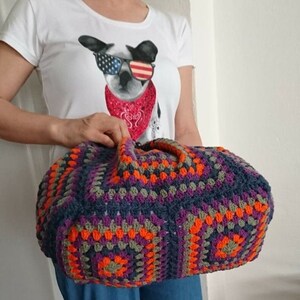 K&ouml;nnte beinhalten: Eine gro&szlig;e, handgefertigte H&auml;keltasche mit geometrischem Muster. Die Tasche hat eine Mischung aus Farben, darunter Marineblau, Orange, Lila und Grau. Sie hat zwei Griffe und eine abgerundete Form. Die Person tr&auml;gt ein wei&szlig;es T-Shirt mit einem Hunde-Motiv.
