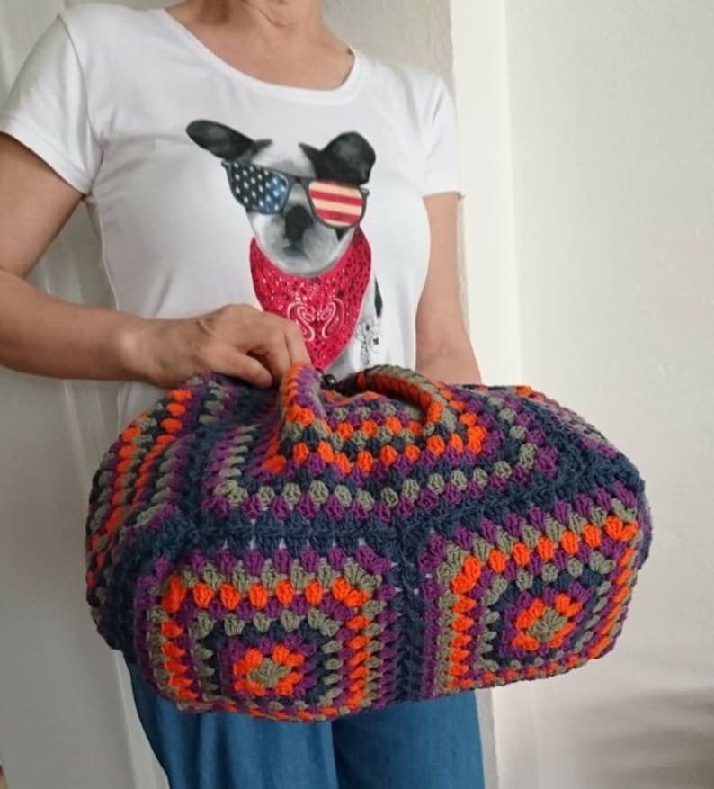 K&ouml;nnte beinhalten: Eine gro&szlig;e, handgefertigte H&auml;keltasche mit geometrischem Muster. Die Tasche hat eine Mischung aus Farben, darunter Marineblau, Orange, Lila und Grau. Sie hat zwei Griffe und eine abgerundete Form. Die Person tr&auml;gt ein wei&szlig;es T-Shirt mit einem Hunde-Motiv.