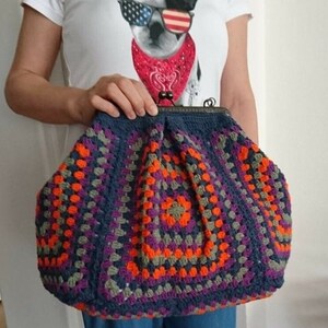 K&ouml;nnte beinhalten: Handgefertigte H&auml;keltasche im Granny-Square-Muster. Die Tasche hat eine dunkelblaue Basis mit orangefarbenen, lila und grauen Quadraten. Sie hat einen Metallverschluss und eine abgerundete Form.