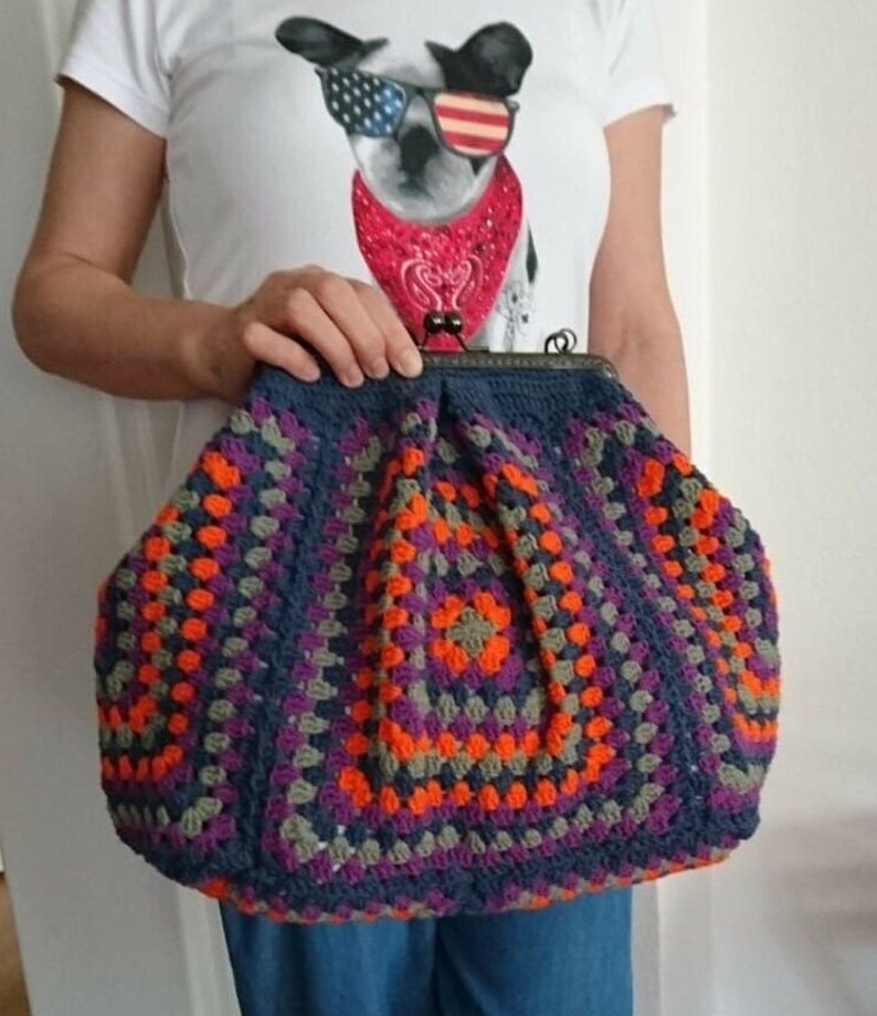 K&ouml;nnte beinhalten: Handgefertigte H&auml;keltasche im Granny-Square-Muster. Die Tasche hat eine dunkelblaue Basis mit orangefarbenen, lila und grauen Quadraten. Sie hat einen Metallverschluss und eine abgerundete Form.