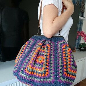 K&ouml;nnte beinhalten: Handgefertigte H&auml;keltasche mit dunkelblauem Grund und einem farbenfrohen Granny-Square-Muster in Orange, Lila und Gr&uuml;n. Die Tasche hat einen braunen Lederriemen und einen Metallverschluss.