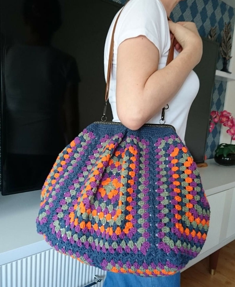 K&ouml;nnte beinhalten: Handgefertigte H&auml;keltasche mit dunkelblauem Grund und einem farbenfrohen Granny-Square-Muster in Orange, Lila und Gr&uuml;n. Die Tasche hat einen braunen Lederriemen und einen Metallverschluss.
