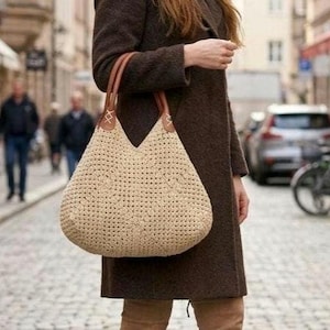 Könnte beinhalten: Beige gehäkelte Tragetasche mit braunen Ledergriffen. Die Tasche hat eine V-förmige Öffnung und wird von einer Person getragen, die einen braunen Mantel trägt. Das gewebte Design der Tasche ist sichtbar, und die Griffe sind mit braunen Nähten befestigt.
