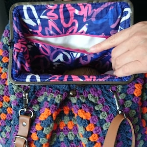 K&ouml;nnte beinhalten: Handgefertigte H&auml;keltasche mit einem farbenfrohen Granny-Square-Muster in Blau-, Orange-, Lila- und Gr&uuml;nt&ouml;nen. Die Tasche hat einen Metallverschluss und einen braunen Schulterriemen aus Leder. Das Innenfutter zeigt ein abstraktes Design in Blau und Rosa.