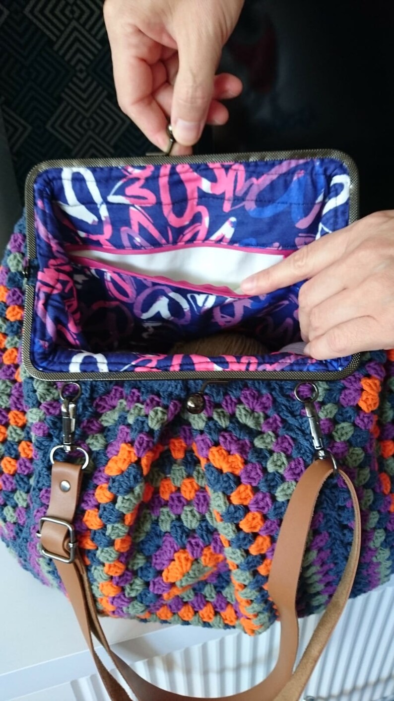 K&ouml;nnte beinhalten: Handgefertigte H&auml;keltasche mit einem farbenfrohen Granny-Square-Muster in Blau-, Orange-, Lila- und Gr&uuml;nt&ouml;nen. Die Tasche hat einen Metallverschluss und einen braunen Schulterriemen aus Leder. Das Innenfutter zeigt ein abstraktes Design in Blau und Rosa.
