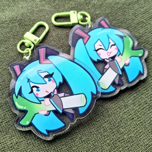 May include: Two acrylic keychains featuring a cartoon character with turquoise hair, pink accents, and a smiling expression. The keychains have a green clasp and are on a green background.