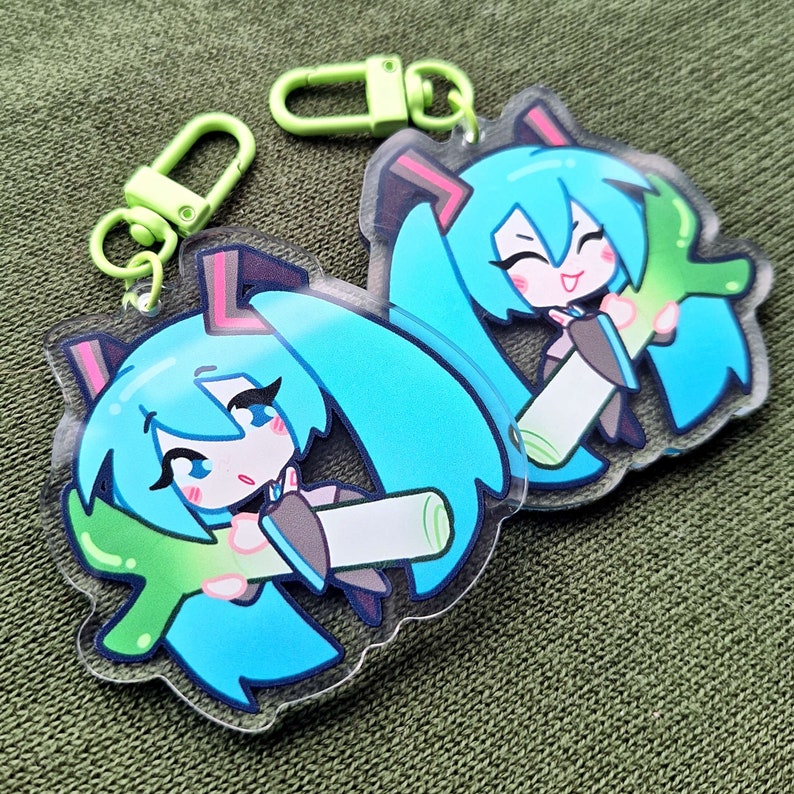 May include: Two acrylic keychains featuring a cartoon character with turquoise hair, pink accents, and a smiling expression. The keychains have a green clasp and are on a green background.