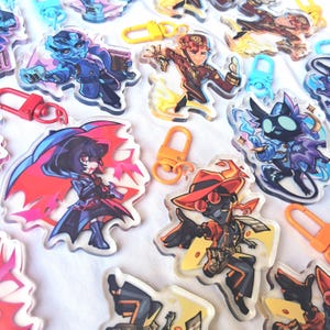 May include: A collection of clear acrylic keychains featuring various illustrated cartoon characters in dynamic poses. Each keychain is attached to a colorful plastic lobster clasp in shades of red, orange, yellow, and light blue, showcasing vibrant artwork.