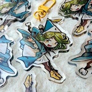 Könnte beinhalten: Transparenter Acryl-Charm mit einer Cartoonfigur mit grünen Haaren, einem blauen Hut und einem blauen Umhang. Die Figur befindet sich in einer dynamischen Pose, mit einem gelben Verschluss. Mehrere Charms sind auf der Oberfläche verstreut.