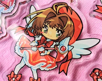 May include: A clear acrylic keychain featuring a chibi-style anime character with brown hair, wearing a red and white dress with a matching red bow. The character holds a staff with a wing design and has small white wings on their back.