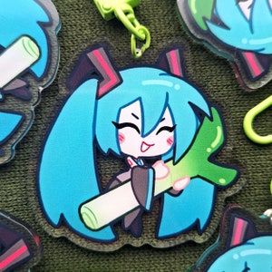 May include: A clear acrylic charm featuring a cartoon character with long, turquoise pigtails and a smiling expression. The character is holding a green and white leek. The charm has a green clasp.
