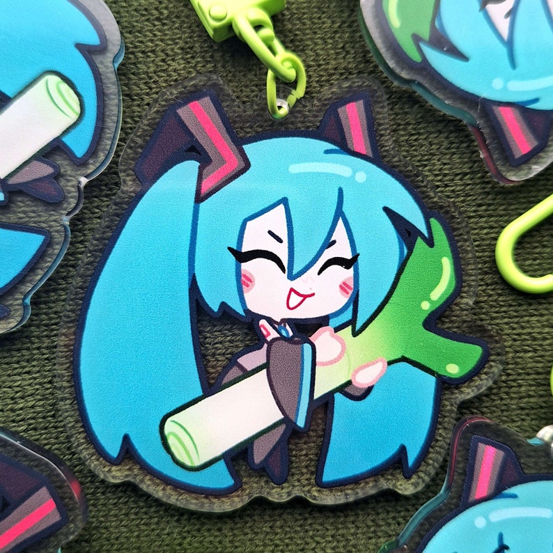 May include: A clear acrylic charm featuring a cartoon character with long, turquoise pigtails and a smiling expression. The character is holding a green and white leek. The charm has a green clasp.