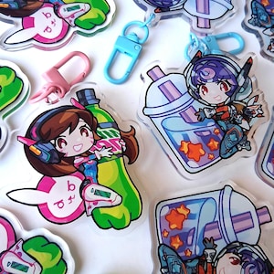 May include: A selection of clear acrylic keyrings featuring anime-style characters. The keyrings depict characters with brown and purple hair, holding a green bottle and a blue drink. The keyrings have pink, blue, and clear accents.