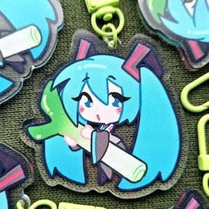 May include: A clear acrylic charm featuring a cartoon character with turquoise hair and a green leek. The charm has a lime green clasp and is surrounded by other similar charms. The character has large blue eyes and is holding a leek.
