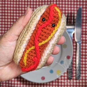 May include: A crocheted hot dog toy with a smiling face. The bun is beige, the sausage is orange, and red and yellow yarn represent ketchup and mustard. Black beads form the eyes. The hot dog is held in a hand.