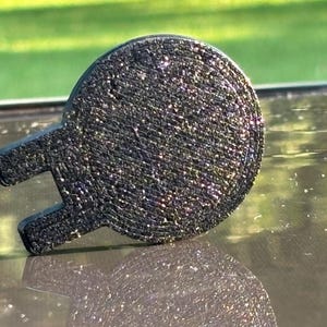 May include: A circular black emblem with a textured, glittery surface and a protruding U-shaped tab on the left side. The object features a dense, granular finish that reflects light, creating a sparkling effect across its entire surface area.