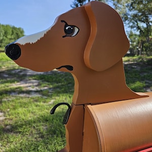 May include: A brown wooden mailbox shaped like a dachshund dog, featuring a painted face with a white snout, black nose, and expressive eyes. The mailbox includes a black metal hook on the side and a red metal flag attached to the side panel.
