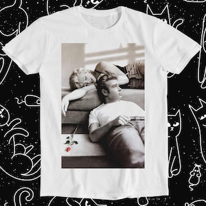May include: A white t-shirt featuring a black and white photograph of a man and woman. The woman is lying down with her head resting on the man's shoulder. The man is sitting up and looking at the camera. A single red rose is on the ground in front of them.