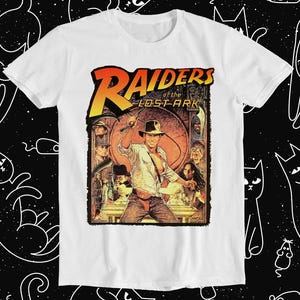 Op de afbeelding: Wit T-shirt met een grafische print van de film Indiana Jones and the Raiders of the Lost Ark. De afbeelding toont Indiana Jones met een zweep in de hand, omringd door andere personages uit de film.