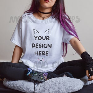 May include: A white t-shirt with the text "YOUR DESIGN HERE" and a cute cat face graphic. The shirt is being worn by a person with purple hair and black fingerless gloves.