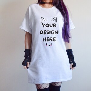 May include: White t-shirt with the text "YOUR DESIGN HERE" in black, featuring cat ears and a smiling face. The shirt is a simple, casual style.