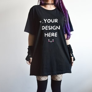May include: Black oversized t-shirt with the text "YOUR DESIGN HERE" in white, along with a cute face graphic. The shirt is worn with black fishnet stockings and arm warmers. The model has purple hair.