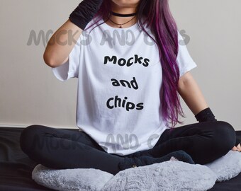 May include: A white t-shirt with the text "Mocks and Chips" in black lettering. The person wearing the shirt has long purple hair and is sitting on a gray and white fuzzy cushion.