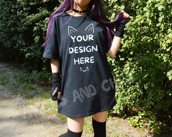 May include: A black oversized t-shirt with the text "YOUR DESIGN HERE" and a cartoon cat face with a smiling mouth. The shirt is being worn by a person with purple hair.