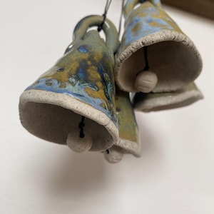 May include: A set of four ceramic bells with a blue, green and brown glaze. The bells are hanging from a string and have a small white bead at the bottom.