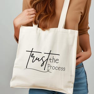 May include: Cream-colored tote bag with the words "Trust the Process" in black script. The bag has two handles and is held by a person. The text is in a cursive font with a small heart symbol below the word "Process".