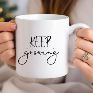 K&ouml;nnte beinhalten: Wei&szlig;e Keramik-Tasse mit dem Schriftzug "KEEP growing" in schwarzer Schrift. Die Tasse hat einen gebogenen Henkel. Die Tasse wird von einer Person gehalten. Die Tasse hat eine Standardgr&ouml;&szlig;e f&uuml;r Kaffee oder Tee.