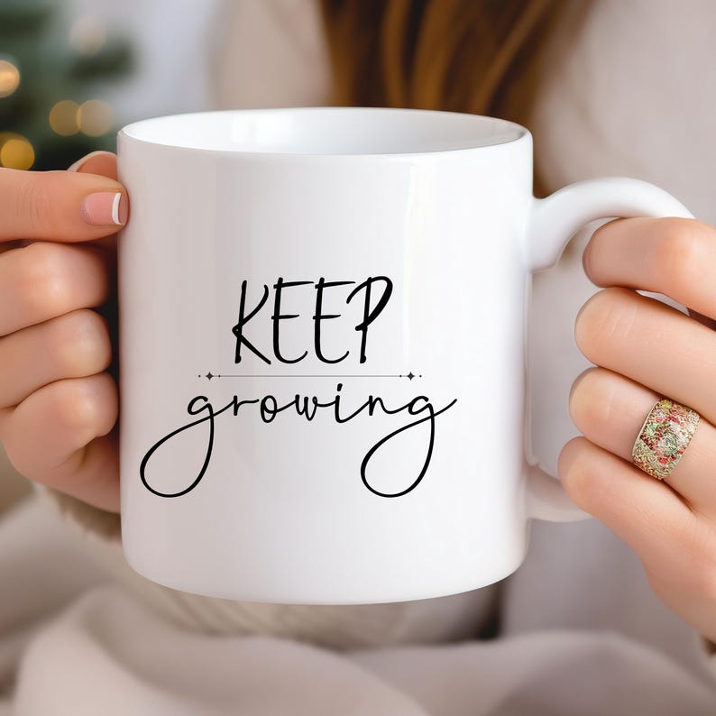 K&ouml;nnte beinhalten: Wei&szlig;e Keramik-Tasse mit dem Schriftzug "KEEP growing" in schwarzer Schrift. Die Tasse hat einen gebogenen Henkel. Die Tasse wird von einer Person gehalten. Die Tasse hat eine Standardgr&ouml;&szlig;e f&uuml;r Kaffee oder Tee.