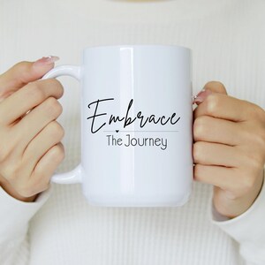 May include: White ceramic mug with the words "Embrace The Journey" in black script. The mug has a large handle and is held by a person wearing a white sweater. The mug is a standard size for coffee or tea.