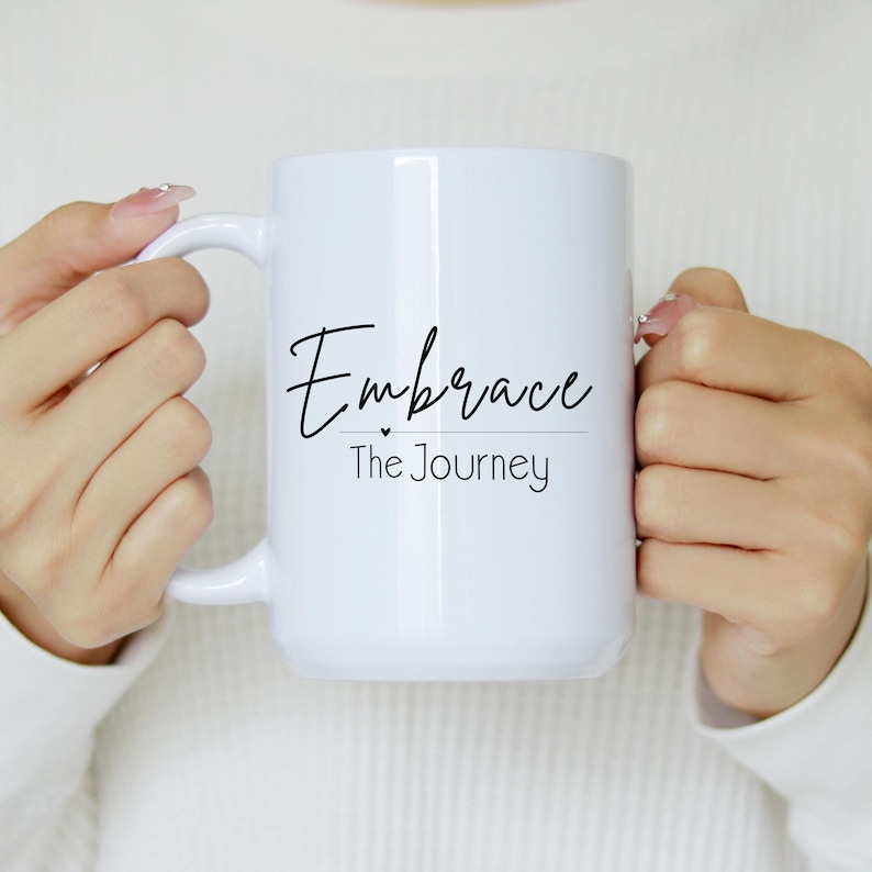 May include: White ceramic mug with the words "Embrace The Journey" in black script. The mug has a large handle and is held by a person wearing a white sweater. The mug is a standard size for coffee or tea.