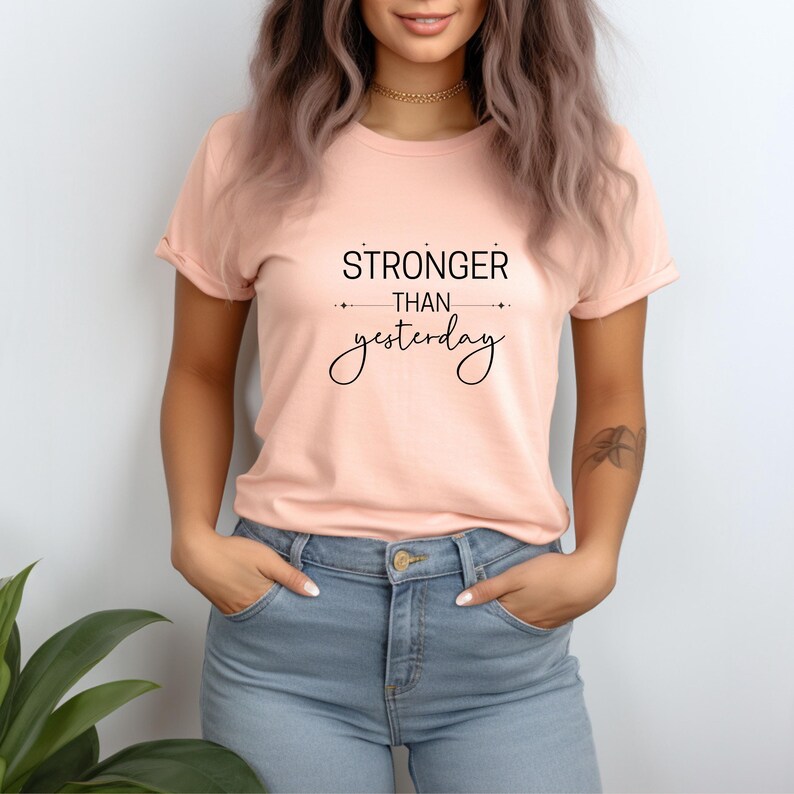 K&ouml;nnte beinhalten: Pfirsichfarbenes T-Shirt mit dem schwarzen Text "STRONGER than yesterday". Das Shirt hat kurze &Auml;rmel und einen Rundhalsausschnitt. Das Model tr&auml;gt eine blaue Jeans.