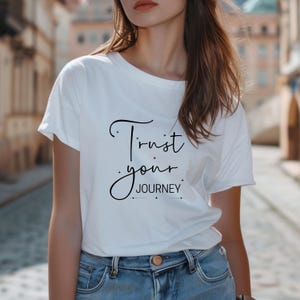 K&ouml;nnte beinhalten: Wei&szlig;es T-Shirt mit dem Text "Trust your JOURNEY" in schwarzer Schrift. Das Shirt hat einen Rundhalsausschnitt und kurze &Auml;rmel. Das Model tr&auml;gt eine blaue Jeans.