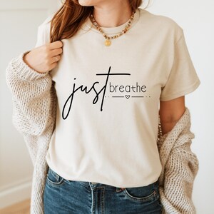 May include: A cream-colored t-shirt with the words "just breathe" in black script. The word "just" is in a flowing script, and "breathe" is in a simple sans-serif font. A small heart symbol is below the word "breathe".