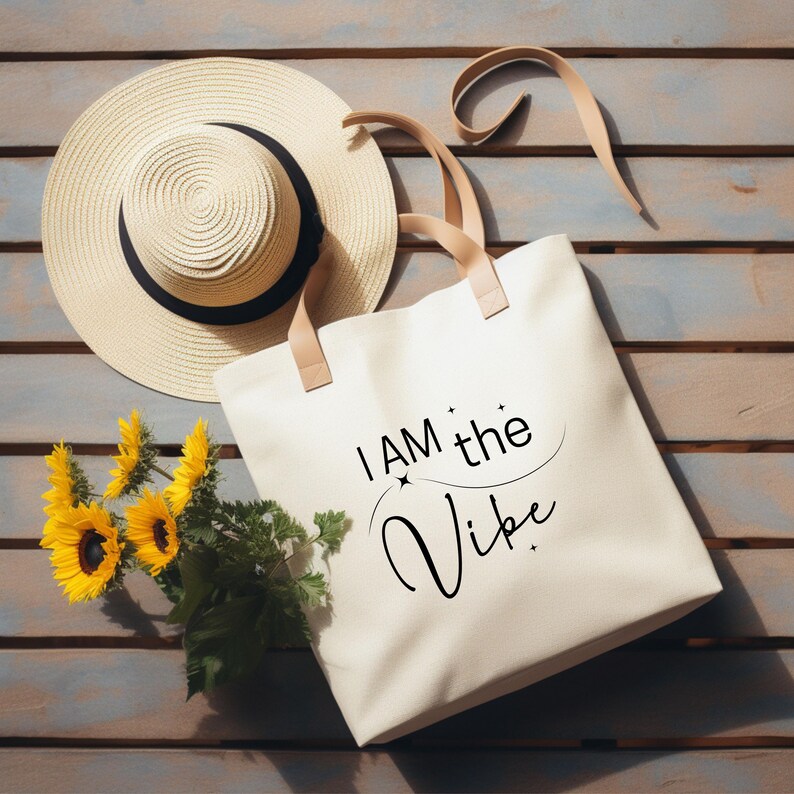 K&ouml;nnte beinhalten: Eine cremefarbene Tragetasche mit dem Text "I AM the Vibe" in schwarzer Schrift, neben einem Sonnenhut aus Stroh mit schwarzem Band und einem Strau&szlig; Sonnenblumen. Die Tasche hat beige Griffe.