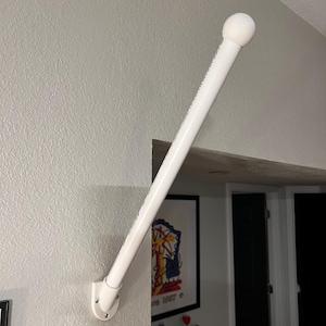 Puede incluir: Una barra de cortina blanca, en ángulo y montada en una pared gris claro texturizada. La barra tiene un remate redondeado en la parte superior y se asegura con un soporte blanco. Diseñada para colgar cortinas.