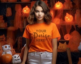 May include: A woman wearing an orange t-shirt with the text "YOUR Design HERE" in brown lettering. She is standing in front of a background of Halloween decorations, including pumpkins and ghosts.