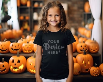 Può includere: Una bambina che indossa una maglietta nera con il testo "YOUR Design HERE" in grigio. È in piedi davanti a uno scaffale con zucche intagliate.
